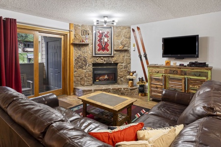 Cozy living room with brown leather sofas, stone fireplace, mounted skis, rustic wooden cabinet, wall-mounted TV, and sliding glass doors leading to a balcony. Room decor includes a small welcome sign.