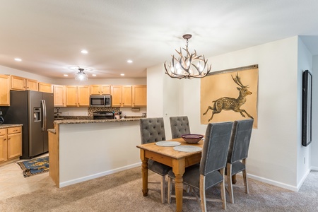 A modern kitchen with wooden cabinets, stainless steel appliances, and a granite countertop is adjacent to a dining area with a wooden table, upholstered chairs, and a chandelier with antler design.