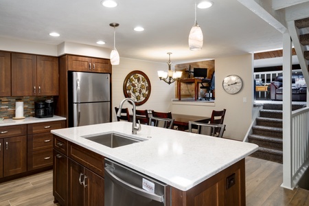 A modern kitchen with dark wooden cabinets, stainless steel appliances, a center island with a sink, and recessed lighting. A dining area with a round table and chairs is in the background near the stairs.