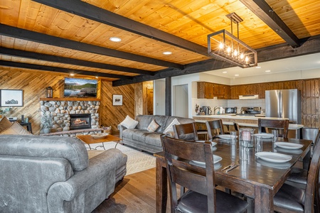 A cozy living and dining area with a rustic design features wooden beams, a stone fireplace, gray sofas, a wooden dining table with six chairs, and an open-concept kitchen with modern appliances.