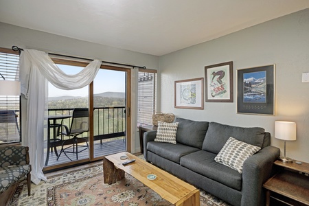 A living room with a dark gray sofa, wooden coffee table, and framed art on the wall. Sliding doors lead to a balcony with a view of a landscape. Pale green walls and patterned rug are visible.