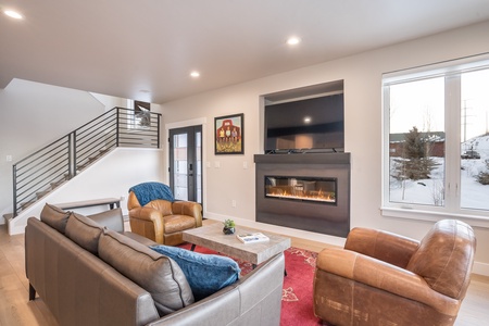 A modern living room with a gray couch, two brown armchairs, a fireplace, a wall-mounted TV, and large windows. A staircase with a black railing is on the left side.