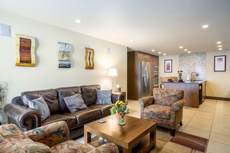 Living room with leather sofa, patterned armchairs, wooden coffee table, and kitchen in the background. Walls feature modern art, and there's a table lamp. Kitchen has stainless steel appliances.