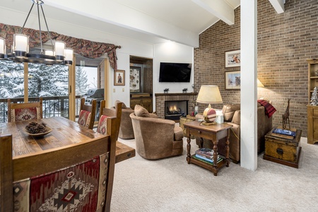 A cozy living room features a brick accent wall, a fireplace, a mounted TV, and comfortable seating. A wooden dining table is in the foreground, and large windows offer a view of a snowy landscape outside.