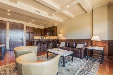 A modern living room with wood paneling features a leather sofa, two upholstered chairs, a large patterned rug, and a coffee table. The background shows a kitchen area with a counter and bar stools.