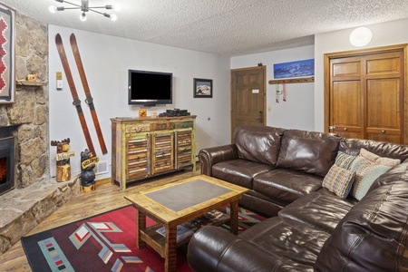 A cozy living room with a leather sectional sofa, wooden coffee table, wooden cabinets, wall-mounted TV, and a stone fireplace. Walls are decorated with skis, a painting, and various decorations.