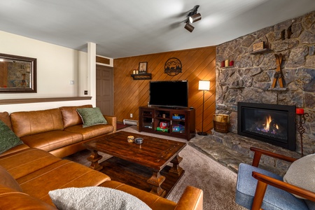 A cozy living room with a leather sectional sofa, wooden coffee table, TV on a stand, stone fireplace, and wood-paneled wall.