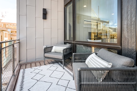 A small outdoor balcony features two gray chairs with striped pillows, a geometric-patterned rug, and glass panel railings. Building construction is visible in the background.