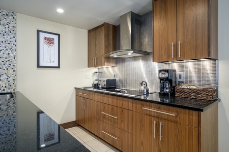 Modern kitchen with wooden cabinets, black granite countertops, a stainless steel exhaust hood, toaster, kettle, coffee maker, and framed wall art. Backsplash has a gray tiled pattern.