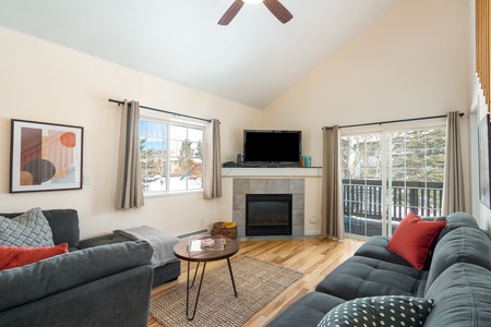 Living room with gray sectional sofas, a wooden coffee table, a fireplace with a TV above, modern decor, large windows, and sliding doors leading to a balcony with tree views.