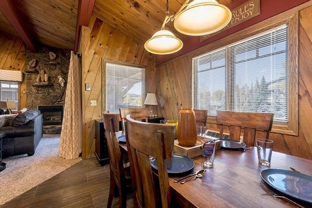 A wooden dining table set for six with plates and glasses is in a room with wood-paneled walls and large windows. A cozy living area with a stone fireplace is visible in the background.