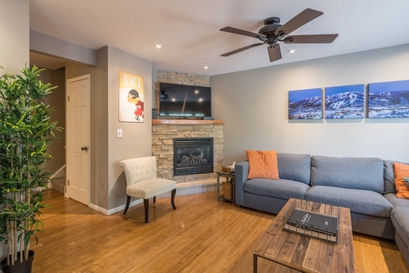 A cozy living room with a couch, coffee table, and fireplace. The wall features a TV above the fireplace, ski-themed artwork, and mountain photos. A ceiling fan and potted plant are also present.