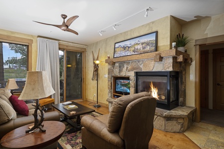 Cozy living room with a stone fireplace, a mounted TV, armchairs, a wooden coffee table, and large windows with curtains. A ceiling fan is above, and a landscape painting hangs over the fireplace.