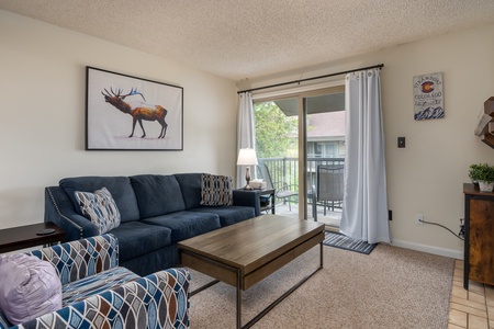 Blue couch, patterned arm chair, coffee table, painting of elk above couch, sliding glass door to porch.