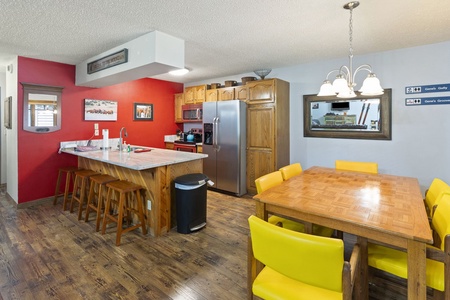 A kitchen with wooden cabinets, a stainless steel refrigerator, a bar with stools, and a dining table with yellow chairs. The floor is wooden, and one wall is painted red while others are light blue.