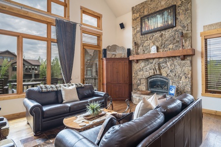 A cozy living room with large windows, two leather couches, a wooden table, and a stone fireplace. A rustic wooden cabinet stands beside the fireplace, and outdoor scenery is visible through the windows.