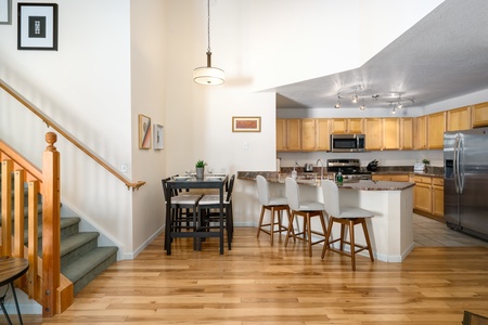 A modern kitchen with light wood cabinets, stainless steel appliances, and a granite island surrounded by four stools. A dining area and staircase with wooden railings are adjacent.