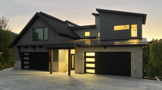 A modern two-story house with dark gray and beige exterior, large windows, and a three-car garage, illuminated at dusk.