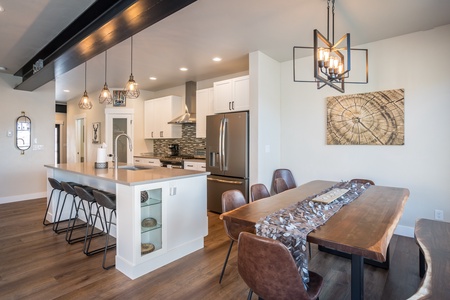 Modern kitchen and dining area with wooden floors, an island with bar stools, contemporary lighting fixtures, stainless steel appliances, and a large wooden dining table.