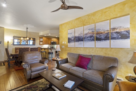 Living room with a brown sofa, two armchairs, a wooden coffee table, and a multi-panel painting on a yellow wall. A kitchen and dining area are visible in the background.