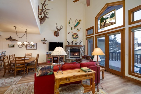 A cozy living and dining area with a fireplace, wooden furniture, and various mounted animal heads and decor on the walls. Large windows provide a view of a snowy outdoor scene.