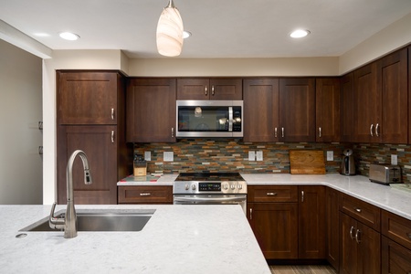 A modern kitchen with dark wood cabinets, stainless steel appliances, a tile backsplash, and a white countertop with an integrated sink. Pendant and recessed lights illuminate the space.