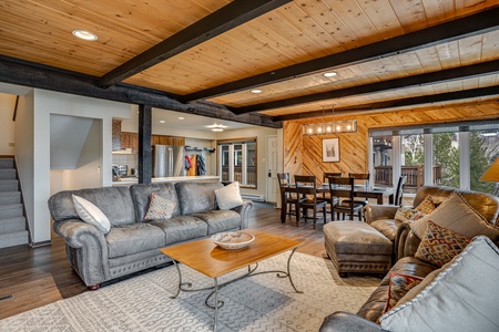 A cozy living room with wooden beams on the ceiling, gray sofas, a wooden coffee table, and a view of the kitchen and dining area. There is a staircase on the left and a large window on the right.