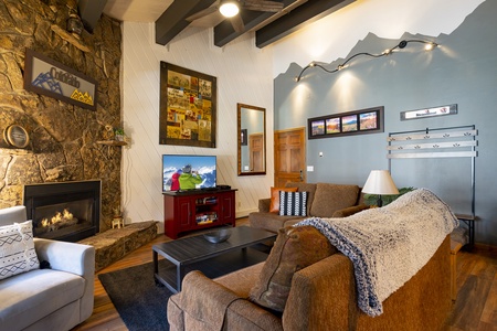 A cozy living room features a stone fireplace, a wall-mounted TV, brown sofas, a white armchair, and a mountain-themed wall decor. The room has wooden floors and warm lighting.