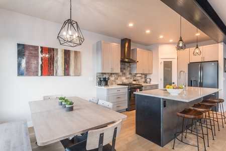 Modern kitchen and dining area with a large table, six chairs, an island with bar stools, pendant lighting, and contemporary decor. Stainless steel appliances and abstract wall art are visible.