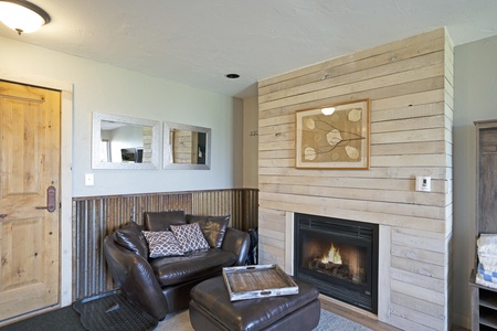 A cozy room with a wooden door, two mirrors, a brown leather chair, ottoman, wooden panel fireplace, and a framed artwork above the fireplace. The room has a rustic decor with a warm atmosphere.