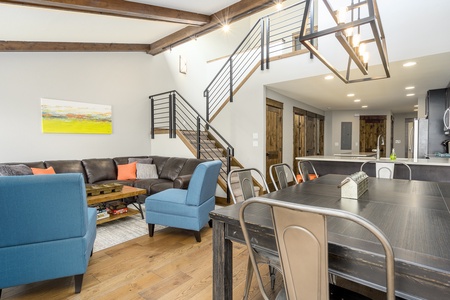 A modern living and dining area features a gray sectional sofa with orange and gray pillows, blue armchairs, a wooden coffee table, metal dining chairs, and a staircase leading to the upper level.