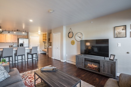 A modern living room with a wall-mounted TV above an electric fireplace, an adjacent kitchen with bar stools, and a clock on the wall. A sofa, coffee table with books, and framed art are also present.