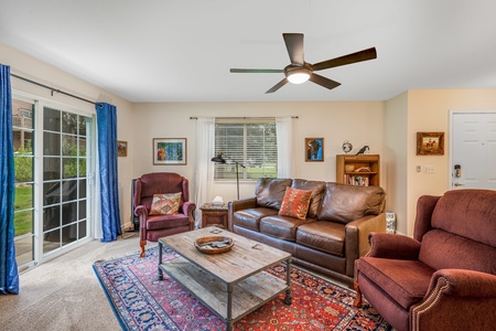 A cozy living room features a brown leather sofa, two armchairs, a wooden coffee table, and a ceiling fan. Large sliding glass doors with blue curtains lead outside. A patterned rug covers the floor.