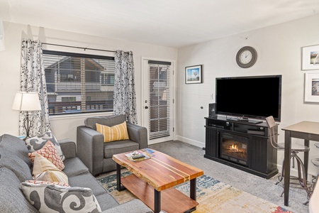 A cozy living room with a gray sofa, armchair, and wooden coffee table. A TV sits on a black stand above a fireplace. The room has a window with curtains, framed pictures, and a door leading outside.