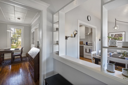 Bright, open kitchen and dining area with white walls, wooden floors, modern light fixtures, and a view into a sunlit room with a table and chairs.