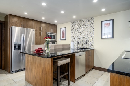 Modern kitchen with wooden cabinets, stainless steel fridge, oven and dishwasher. An island features a vase with flowers and two place settings. Walls are adorned with artwork and mosaic tiles.