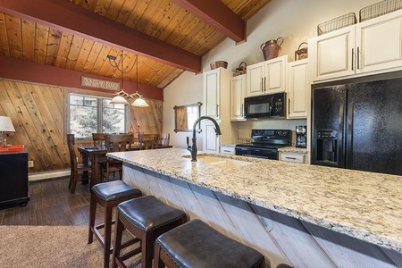 A kitchen and dining area featuring a long granite countertop with barstools, white cabinets, black appliances, a wood-paneled wall, and a wooden dining table with chairs beneath hanging lights.