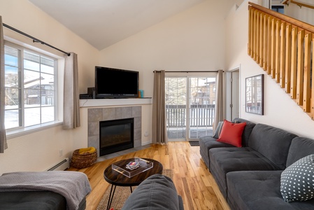 A living room with a gray sofa, red throw pillow, and wooden coffee table facing a TV on a mantel above a fireplace. Windows on the left and sliding glass doors on the right let in natural light.