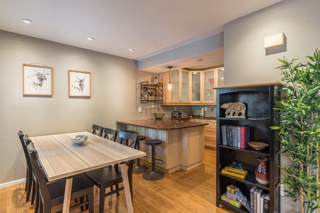 A modern dining area with a wooden table and black chairs, adjacent to a kitchen with a breakfast bar. There are framed pictures on the wall, and a bookshelf with various items is on the right.