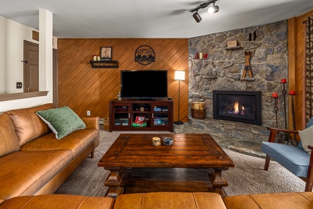 Cozy living room with leather sofas, wooden coffee table, TV on a stand, and a stone fireplace. Walls feature wooden paneling and various decorations, including a wall sign and candles.