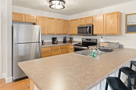 Modern kitchen with stainless steel appliances, light wooden cabinets, beige countertops, and a breakfast bar. Various small appliances and kitchen utensils are placed on the counters.