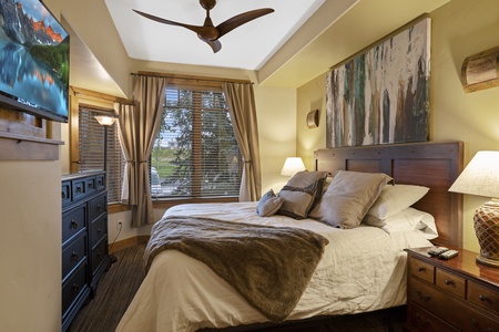 A cozy bedroom with a large bed, wooden furniture, a television mounted on the wall, and a window with a scenic view. The room is well-lit with bedside lamps and a ceiling fan overhead.