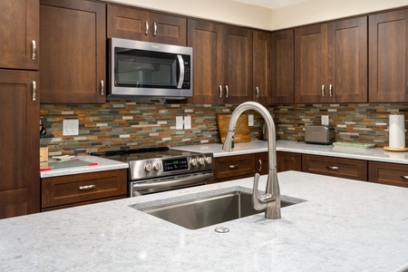 Modern kitchen with wooden cabinets, stainless steel appliances, and a colorful tile backsplash. A central island features a sink with a curved faucet.