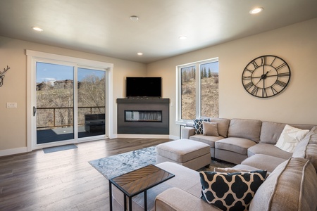 A modern living room with a large sectional sofa, wall-mounted TV above a fireplace, a coffee table, a sliding glass door opening to a balcony, and a wall clock. The room has light walls and wood flooring.