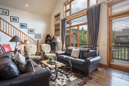 A living room with leather sofas, chairs, and a coffee table on a patterned rug. Large windows offer a view of greenery and houses outside. Stairs and wall art are visible on the left.