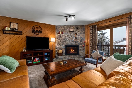 A cozy living room features a stone fireplace, a wooden coffee table, a TV, and seating with green and beige cushions. A large window offers a view of snow-covered trees and a balcony outside.