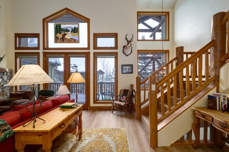 A cozy living room with wooden furniture, a red sofa, a coffee table, two lamps, a staircase, large windows, and a mounted deer head. Snowy outdoor scenery is visible through the windows.