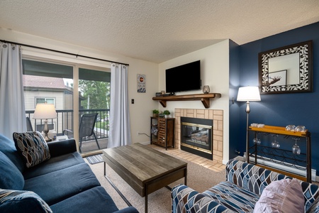 Living Area with blue couch, patterned arm chair, coffee table, gas fireplace with flat screen television above it, sliding glass door with porch.