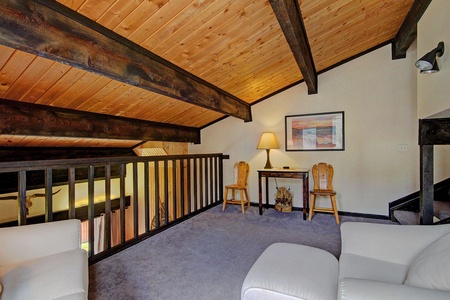 A cozy loft area with wooden ceiling beams, a small desk with a lamp, two wooden chairs, and a framed picture on the wall. The space includes a carpeted floor and white cushioned seating.