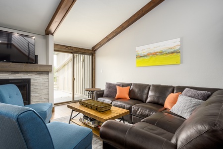 Living room with a brown leather sectional sofa, blue armchairs, a wooden coffee table, wall-mounted TV, fireplace, and sliding glass doors leading to an outdoor area. Bright abstract painting on wall.